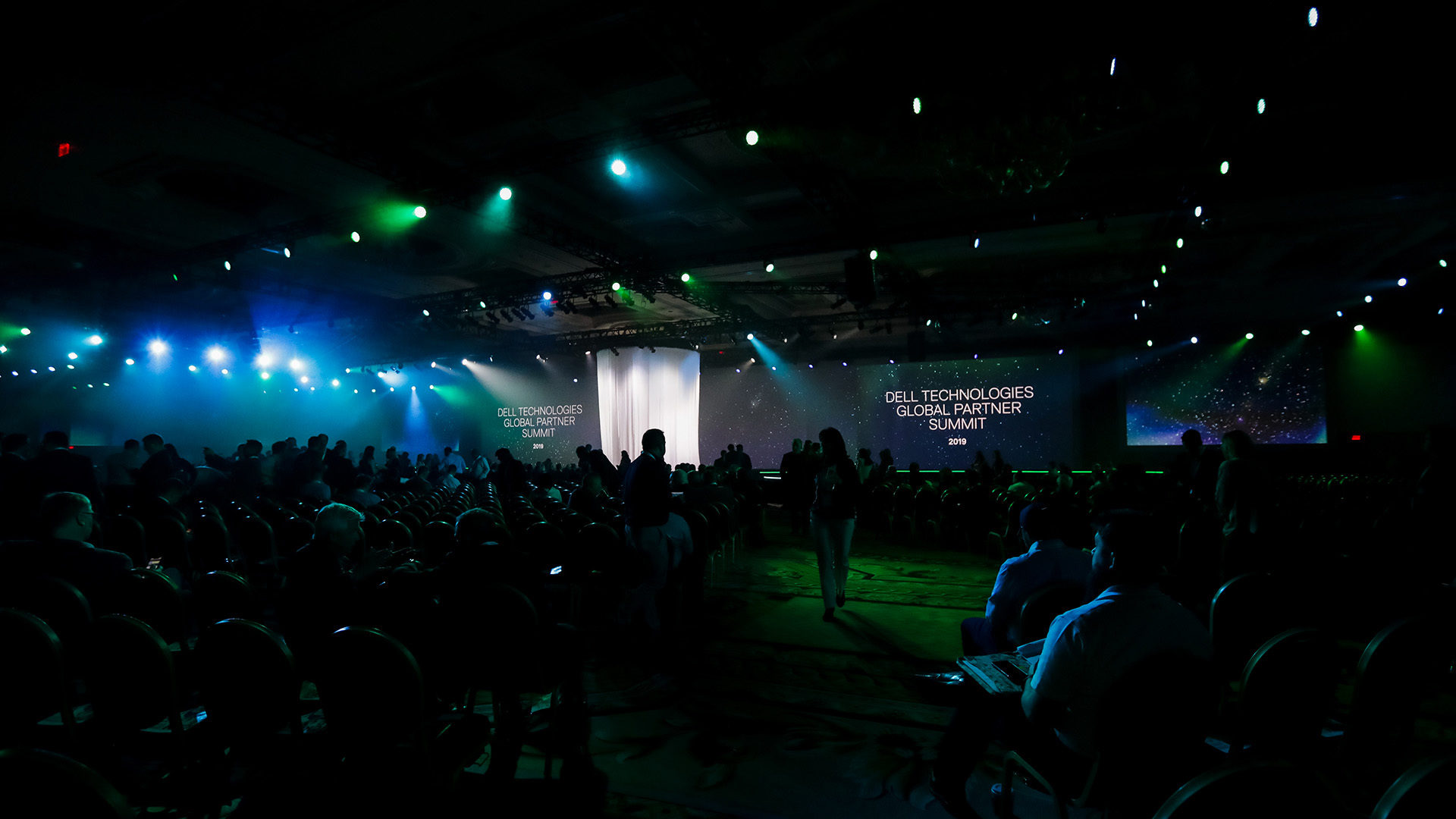 View of the main stage from the back of the room with a full audience and green and blue lights throughout the room.