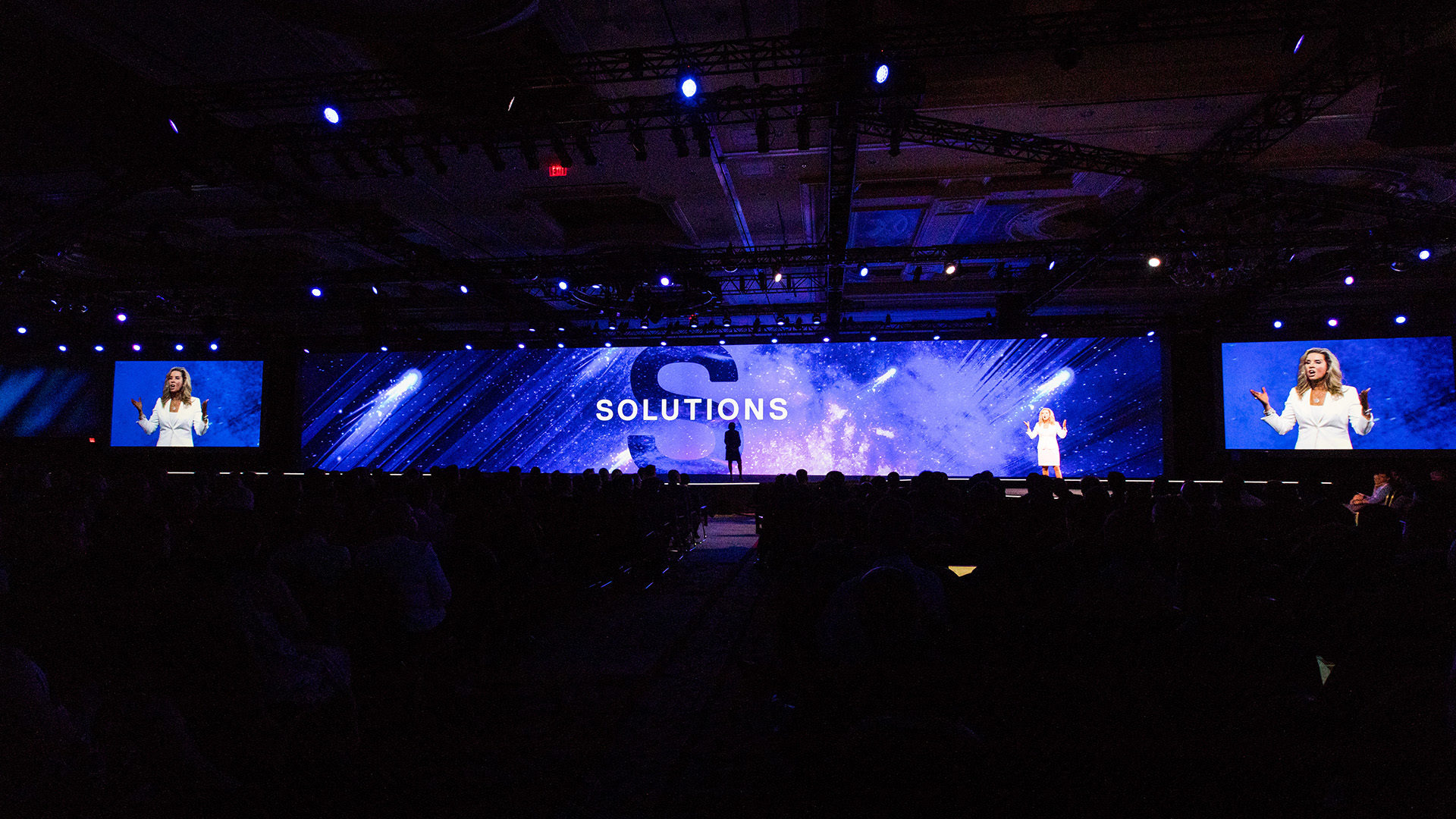 View of the main stage from the back of the room. The woman is speaking in front of a large screen with graphics that read "solutions."