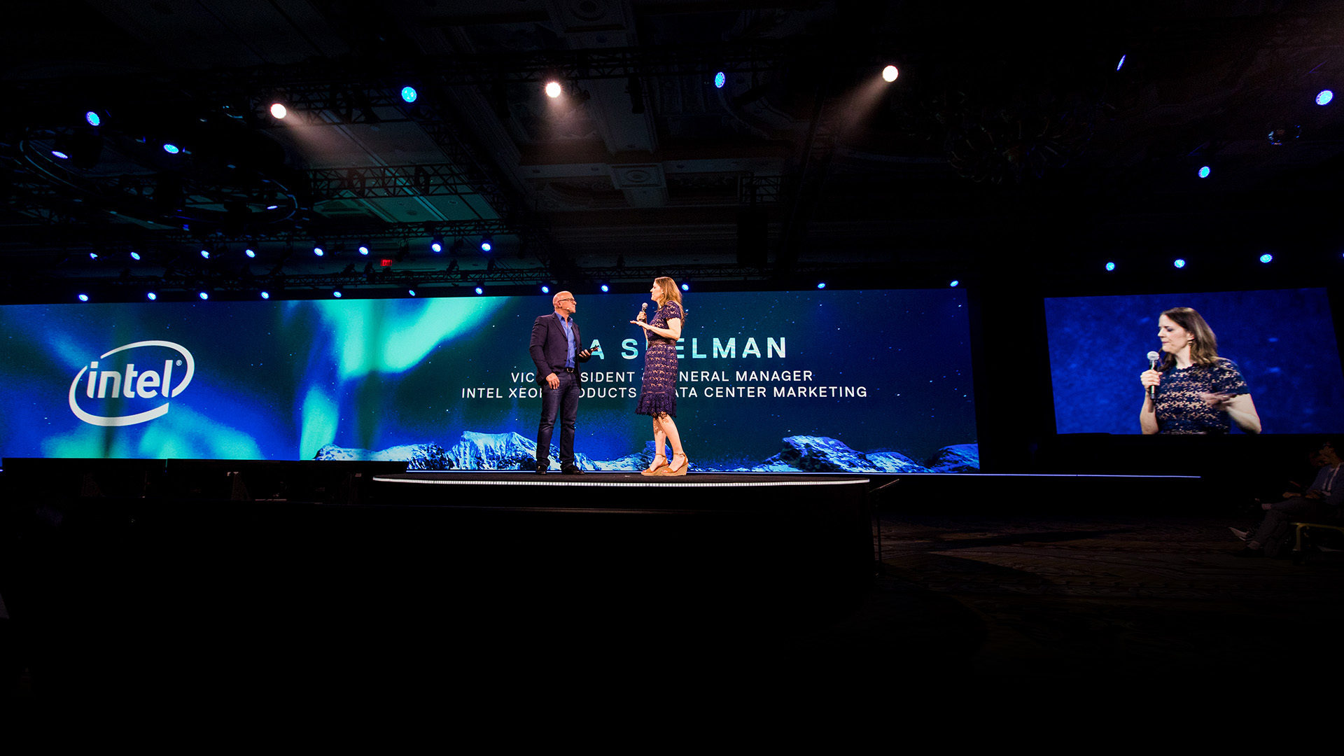 Two people speaking on the main stage in front of the large screen with graphics behind them.